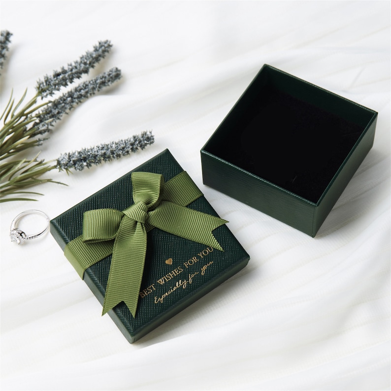 May include: A square dark green jewellery gift box with a textured finish, topped with an olive green ribbon bow. The lid features gold text reading BEST WISHES FOR YOU and Especially for you. The box is open, revealing a black interior lining.