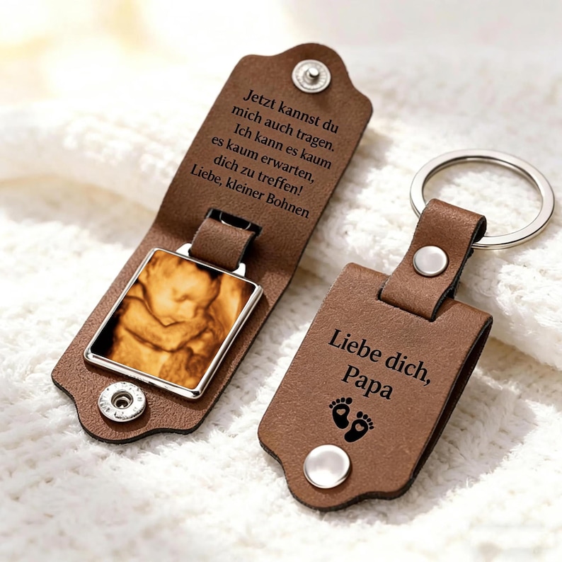 May include: Two brown leather keychains with silver metal rings and snap closures. One keychain is open, revealing a square frame containing a sepia-toned ultrasound image and German text. The other is closed, featuring the text "Liebe dich, Papa" and footprints.