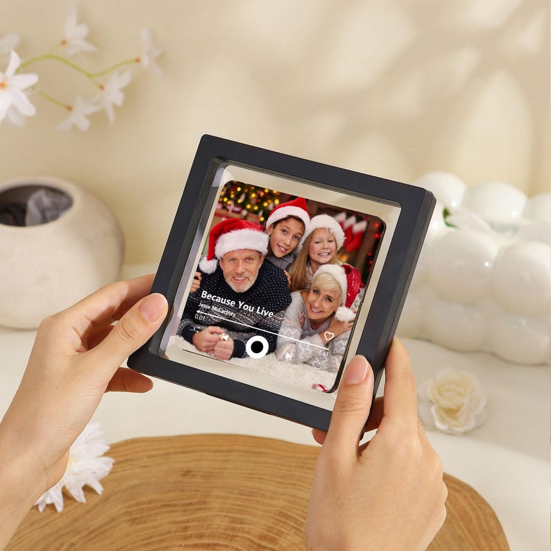 K&ouml;nnte beinhalten: Ein quadratischer, schwarzer schwebender Bilderrahmen mit einem Foto von drei Personen, die Weihnachtsm&uuml;tzen tragen. Das Foto zeigt eine Musik-Player-Oberfl&auml;che mit dem Text Because You Live von Jesse McCartney, einem Fortschrittsbalken.