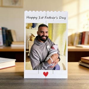 May include: A white photo block with the text "Happy 1st Father's Day" and a red heart. The block displays a photo of a man holding a baby. The man is wearing a gray hoodie.