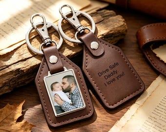 Puede incluir: Dos llaveros de cuero marrón con herrajes de metal plateado. Uno tiene un inserto fotográfico rectangular que muestra a una persona sosteniendo a un bebé. El otro está grabado con el texto: Drive Safe Daddy. I love you! Ambos tienen una anilla.