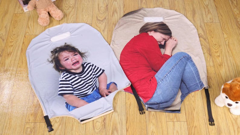 May include: Two gray, padded mats with black straps and buckles. One mat features a crying child in a black and white striped shirt and blue jeans. The other mat shows a person in a red shirt and blue jeans.