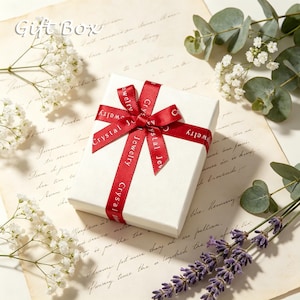 May include: A square white gift box tied with a red satin ribbon featuring the repeating white text Crystal Jewelry. The box is positioned on a piece of vintage-style parchment paper decorated with sprigs of white baby's breath and lavender.