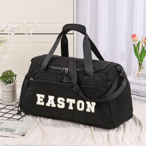 Peut inclure: Sac de sport noir avec le nom "EASTON" en blanc, souligné d'or. Le sac possède une poignée supérieure, une bandoulière et plusieurs compartiments zippés. Une petite poche en filet est visible sur le côté.