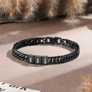 Puede incluir: Una pulsera de eslabones de metal negro con tres cuentas rectangulares grabadas con los nombres Carter, Daniel y Alvaro. La pulsera incluye un mecanismo de cierre seguro y un acabado pulido, creando una apariencia elegante y uniforme.