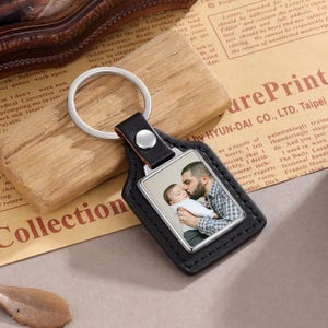 May include: A black leather keyring with a silver ring and a rectangular photo insert. The photo shows a man kissing a baby. The keyring is on a wooden surface with printed text in the background.