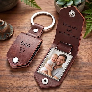 May include: A brown leather keychain with a metal ring and snap closure. The exterior is engraved with the word DAD and a heart symbol. The interior features a photograph of a man holding a baby and the engraved text Drive safe dad, we love you x.