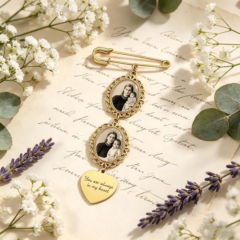 May include: A gold-toned safety pin brooch featuring two oval-shaped frames containing black and white photographs of people, suspended above a heart-shaped charm engraved with the text, You are always in my heart, in a script font.