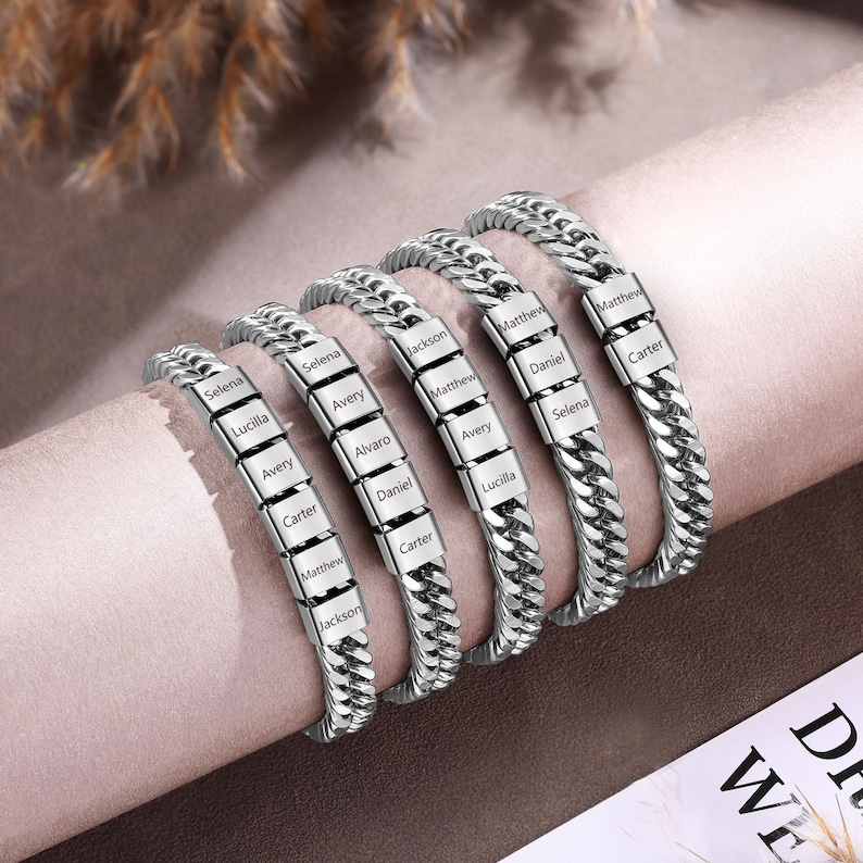 Puede incluir: Cinco pulseras de cadena tipo barbada en tono plateado con cuentas met&aacute;licas rectangulares grabadas con varios nombres, incluyendo Selena, Lucilla, Avery, Carter, Matthew, Jackson, Alvaro y Daniel. Las pulseras est&aacute;n dispuestas en fila sobre un soporte cil&iacute;ndrico.