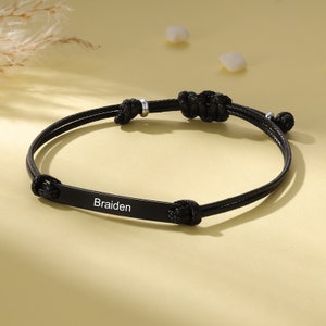 Op de afbeelding: Zwarte verstelbare armband met een zwarte metalen plaat gegraveerd met de naam "Braiden".