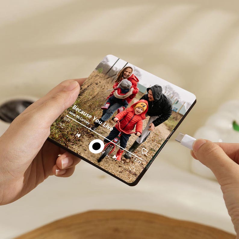 K&ouml;nnte beinhalten: Eine quadratische Acrylplatte mit einem aufgedruckten Foto einer Familie im Freien, &uuml;berlagert mit einer Musik-Player-Oberfl&auml;che, die den Songtitel Because You Live von Jesse McCartney anzeigt, mit einem USB-C-Anschluss an der Seite.