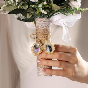 May include: A gold-toned metal safety pin charm featuring two oval-shaped frames with scalloped edges. Each frame contains a small photograph. The charm is attached to a bouquet handle wrapped in textured brown burlap and white lace trim.