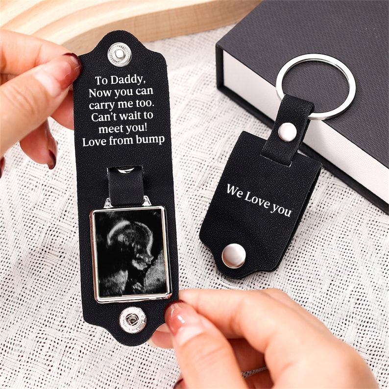 May include: A black leather keychain with a silver metal ring and snap buttons. The interior features the text: To Daddy, Now you can carry me too. Can't wait to meet you! Love from bump. A second folded keychain displays the text: We Love you.
