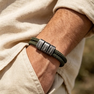 May include: A personalized bracelet with a dark green rope band. The bracelet features three silver-colored cylindrical beads engraved with the names "Loana", "Martin", and "Fabian". The bracelet is worn on a wrist.