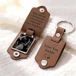 May include: Two brown leather keychains with silver metal rings and snap closures. One keychain is open, revealing a black and white ultrasound image and text reading: Now you can carry me too. Can't wait to meet you! Love, little Bean. The other is closed, displaying: Love You Daddy with a heart and footprint icon.