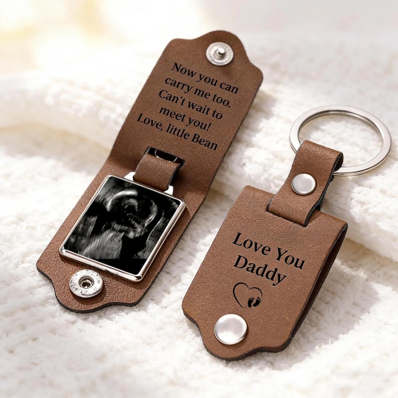 May include: Two brown leather keychains with silver metal rings and snap closures. One keychain is open, revealing a black and white ultrasound image and text reading: Now you can carry me too. Can't wait to meet you! Love, little Bean. The other is closed, displaying: Love You Daddy with a heart and footprint icon.