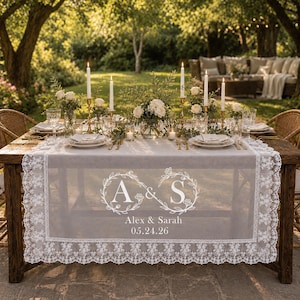 May include: A white sheer table runner with intricate lace trim edges, featuring a printed design of an infinity symbol formed by floral wreaths surrounding the letters A and S, with the text Alex & Sarah and the date 05.24.26 centered below.