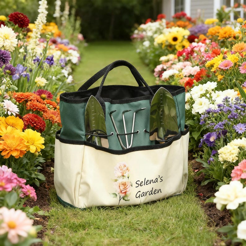 Pu&ograve; includere: Una borsa da giardinaggio verde e crema con manici e finiture nere, contenente attrezzi da giardinaggio. La borsa presenta la scritta "Selena's Garden" e un motivo floreale. La borsa &egrave; posizionata su un sentiero erboso fiancheggiato da fiori colorati.