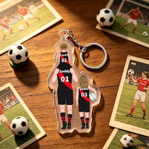 May include: A clear acrylic keychain featuring a printed illustration of a person carrying a child on their shoulders and standing next to another child. The figures wear black and red sports jerseys labeled Jack 02, Daddy 01, and Emma 03.