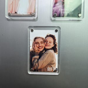 Puede incluir: Un marco de fotos magn&eacute;tico de acr&iacute;lico transparente que contiene una fotograf&iacute;a vertical de dos personas abrazadas y sonriendo. El marco est&aacute; sujeto por cuatro imanes circulares plateados en las esquinas. El texto BFF est&aacute; impreso en blanco abajo a la derecha.
