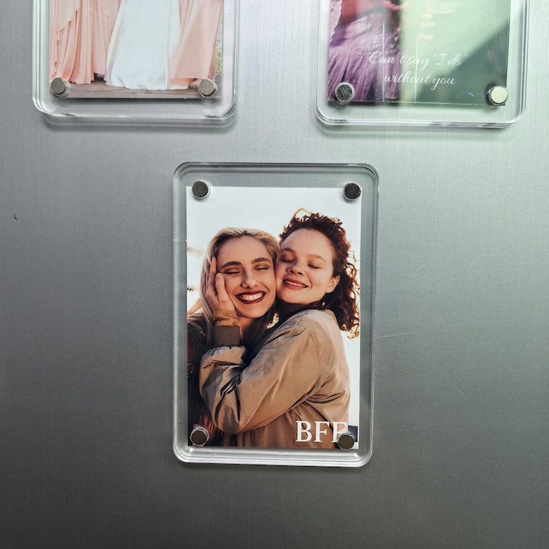 Puede incluir: Un marco de fotos magn&eacute;tico de acr&iacute;lico transparente que contiene una fotograf&iacute;a vertical de dos personas abrazadas y sonriendo. El marco est&aacute; sujeto por cuatro imanes circulares plateados en las esquinas. El texto BFF est&aacute; impreso en blanco abajo a la derecha.