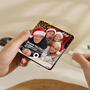 K&ouml;nnte beinhalten: Eine quadratische Acrylplatte mit einem aufgedruckten Foto einer Familie mit Weihnachtsm&uuml;tzen. Das Bild enth&auml;lt eine Musik-Player-Oberfl&auml;che mit dem Text Because You Live von Jesse McCartney, einer Wiedergabetaste und einem Herzsymbol.
