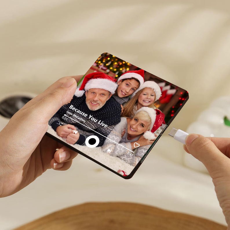 K&ouml;nnte beinhalten: Eine quadratische Acrylplatte mit einem aufgedruckten Foto einer Familie mit Weihnachtsm&uuml;tzen. Das Bild enth&auml;lt eine Musik-Player-Oberfl&auml;che mit dem Text Because You Live von Jesse McCartney, einer Wiedergabetaste und einem Herzsymbol.