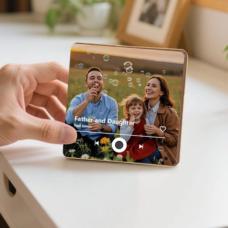 K&ouml;nnte beinhalten: Eine quadratische Acrylplatte mit einem aufgedruckten Foto eines Mannes und eines jungen M&auml;dchens auf einem Feld mit Seifenblasen. Das Design enth&auml;lt eine Musik-Player-Oberfl&auml;che mit dem Text Father and Daughter von Paul Simon und Wiedergabesymbolen.