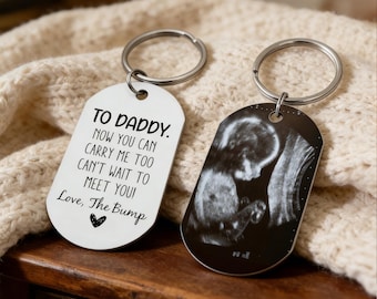 Könnte beinhalten: Zwei silberne Schlüsselanhänger. Einer hat den Text "TO DADDY. NOW YOU CAN CARRY ME TOO CAN'T WAIT TO MEET YOU! Love, The Bump" und ein Herz. Der andere zeigt ein schwarz-weißes Ultraschallbild.