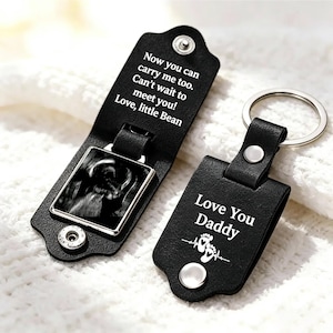 May include: Two black leather keychains with silver metal rings. One is open, revealing a black and white ultrasound image and the text: Now you can carry me too. Can't wait to meet you! Love, little Bean. The other is closed, reading: Love You Daddy.