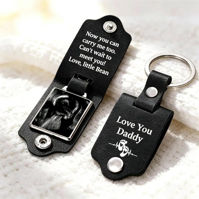 May include: Two black leather keychains with silver metal rings. One is open, revealing a black and white ultrasound image and the text: Now you can carry me too. Can't wait to meet you! Love, little Bean. The other is closed, reading: Love You Daddy.