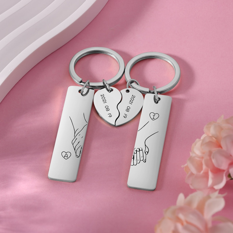 May include: Two silver keychains with matching engraved designs. Each keychain features a hand holding a heart with the text "I" and "A" on the hearts. The keychains also have a heart-shaped charm with the date "2021 08 19" engraved on it.