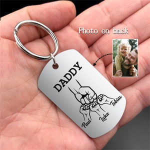 May include: Silver dog tag keyring with the word "DADDY" in black. The tag features a graphic of a fist bump with three smaller fists below, with the names "Paul", "Luka", and "Tobias". A small photo is on the back.