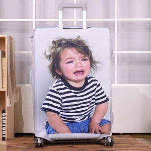 May include: A white suitcase cover featuring a crying child wearing a black and white striped shirt and blue jeans. The suitcase has a retractable handle and wheels. The background includes a bookshelf, a teddy bear, and a toy dog.