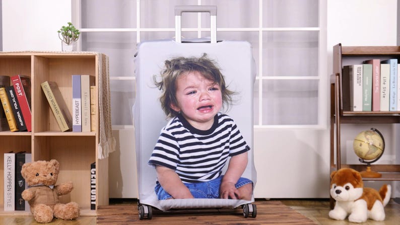 May include: A white suitcase cover featuring a crying child wearing a black and white striped shirt and blue jeans. The suitcase has a retractable handle and wheels. The background includes a bookshelf, a teddy bear, and a toy dog.