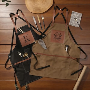 May include: Two aprons, one black and one brown, with leather straps and pockets. The black apron has the name "Kevin" on a leather patch, and the brown apron has the name "Dagmar". Kitchen utensils and silverware are arranged around the aprons.