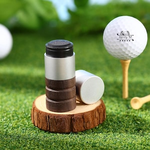May include: A golf-themed image featuring a white golf ball on a wooden tee, alongside a cylindrical golf ball marker with a black top, silver body, and wooden base. The marker is resting on a small wooden slice.