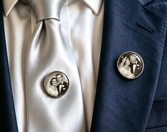 May include: A silver tie and a dark blue blazer featuring two circular metal pins. Each pin displays a black and white photograph of a couple in wedding attire. The tie is solid silver with a smooth finish, worn over a crisp white collared shirt.