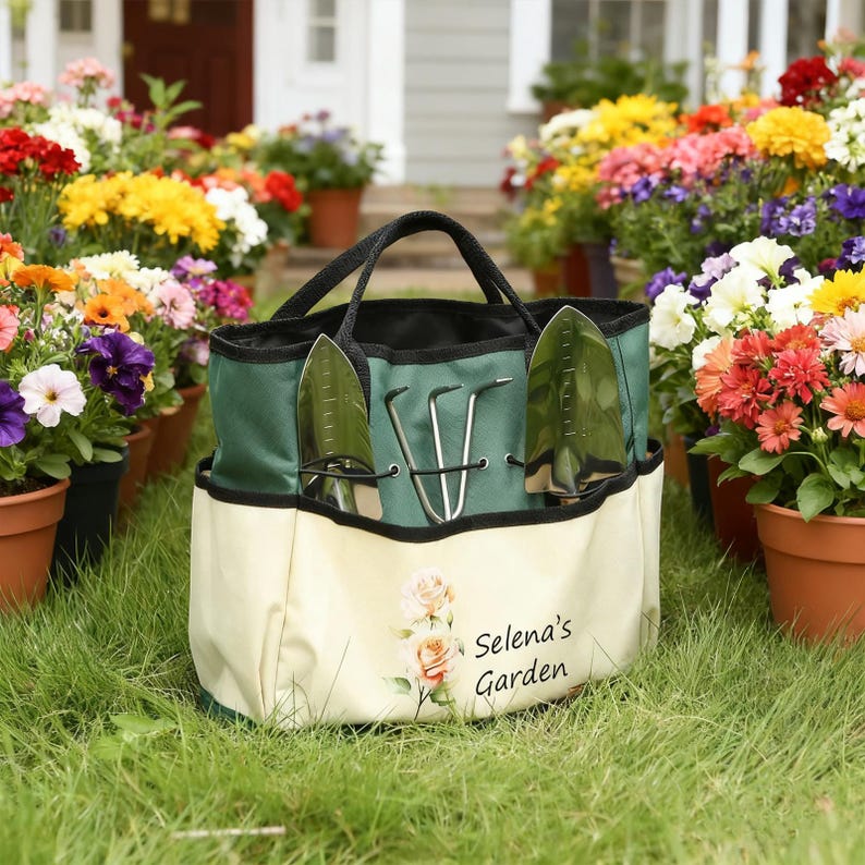Pu&ograve; includere: Una borsa da giardinaggio verde e crema con manici neri e un design floreale. La borsa contiene attrezzi da giardinaggio, tra cui una cazzuola e un rastrello a mano. Il testo "Selena's Garden" &egrave; stampato sul davanti. Fiori in vaso circondano la borsa.