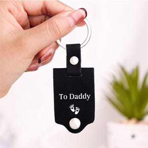 May include: A black rectangular leather keychain featuring a silver metal ring at the top. The front surface is embossed with the white text To Daddy above a graphic of two small baby footprints with heart-shaped heels and a silver snap button.