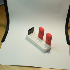 May include: A white 3D-printed organizer holding a black SD card and two red button-shaped devices. The organizer has multiple slots. The items are on a white surface, with a wooden table in the background.