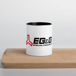 Könnte beinhalten: Weißer Keramikbecher mit schwarzem Innenraum und dem Text "EG&G SPECIAL PROJECTS" in Schwarz und Rot. Der Becher steht auf einem Holzschneidebrett.