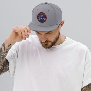 May include: A gray baseball cap with a flat brim. The cap features a circular patch with text and a design, including the words "Joint Strike Fighter" and "United Kingdom Air Force". The cap is worn by a person.