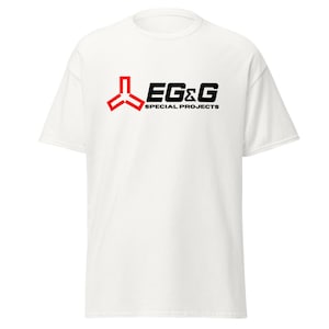 Puede incluir: Camiseta blanca con un diseño rojo y negro. El diseño incluye un símbolo estilizado rojo y el texto "EG&G SPECIAL PROJECTS" en negro. La camiseta está hecha de un material suave y tiene mangas cortas.