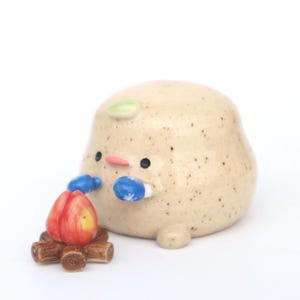 May include: A ceramic figurine of a round, beige creature with black eyes, a pink nose, and a green leaf on its head. It wears blue mittens with snowflake designs and sits next to a miniature campfire with red and yellow flames.