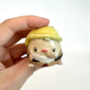 May include: A ceramic gnome figurine with a yellow hat, white beard, and a red nose. The gnome has black eyes and is wearing a brown outfit with black accents. The figurine is small and detailed.