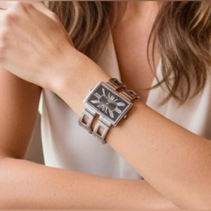 May include: A rectangular wristwatch with a black face and silver hands and markers. The watch has a silver metal band with a unique geometric design. The watch is worn on a wrist.