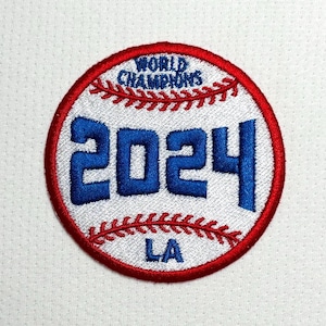 以下が含まれることがあります： 白と赤の野球ボールの形をしたパッチで、「WORLD CHAMPIONS」と「2024」が青色の文字で書かれています。パッチには中央に「LA」の文字もあります。