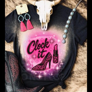 May include: Black t-shirt with a pink and white design featuring the text "Clock it", a high heel, and lipstick. The shirt is accessorized with a turquoise necklace, earrings, a cow skull, and a pair of black cowboy boots.