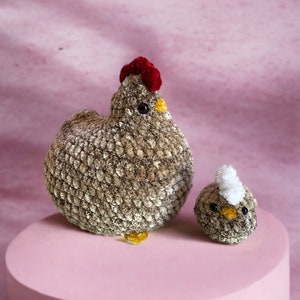 May include: A brown and grey plush chicken with a red comb and yellow beak, and a smaller brown and grey plush chick with a white tuft of feathers on its head, both sitting on a pink circular platform.